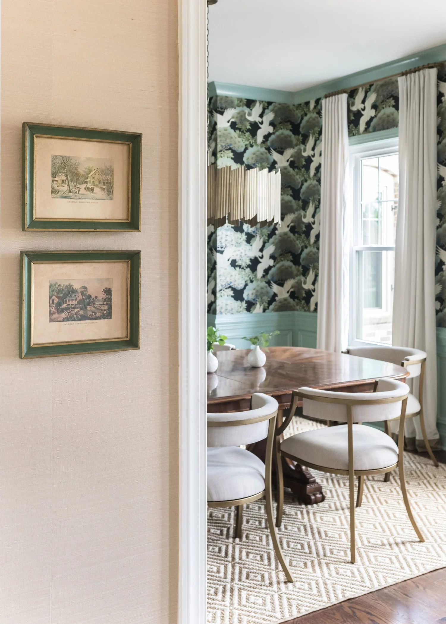 View from a grasscloth hallway into a dining room with dark crane wallpaper, an oval mahogany table and brass chairs