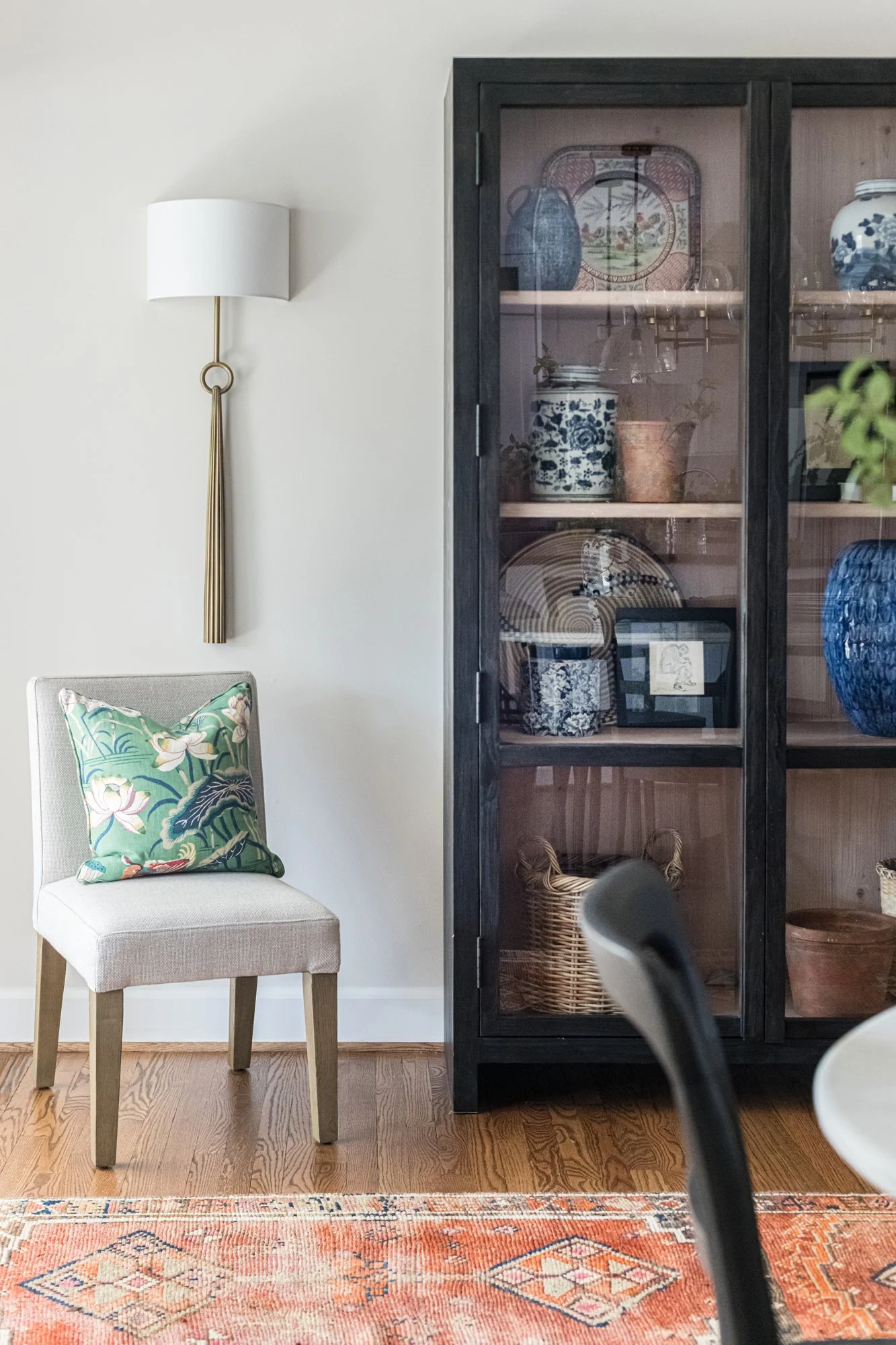 Black glass-front cabinet filled with blue-and-white ceramics beside a pale linen chair on warm oak flooring