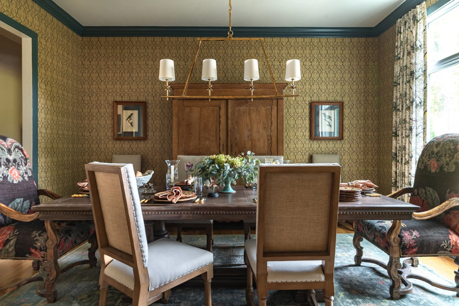 Dark wood trestle dining table set for a meal beneath a brass chandelier, framed by ochre patterned wallpaper